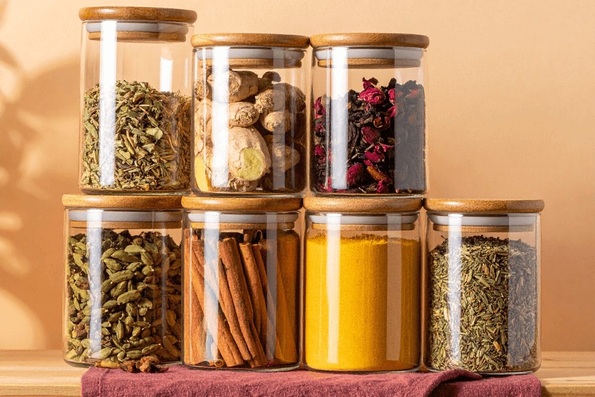 Glass and wooden jars filled with Ayurvedic herbs and spices — ginger, turmeric, fennel, and rose — displayed in a warm saffron and burgundy kitchen setting, symbolizing herbal alchemy and everyday healing.