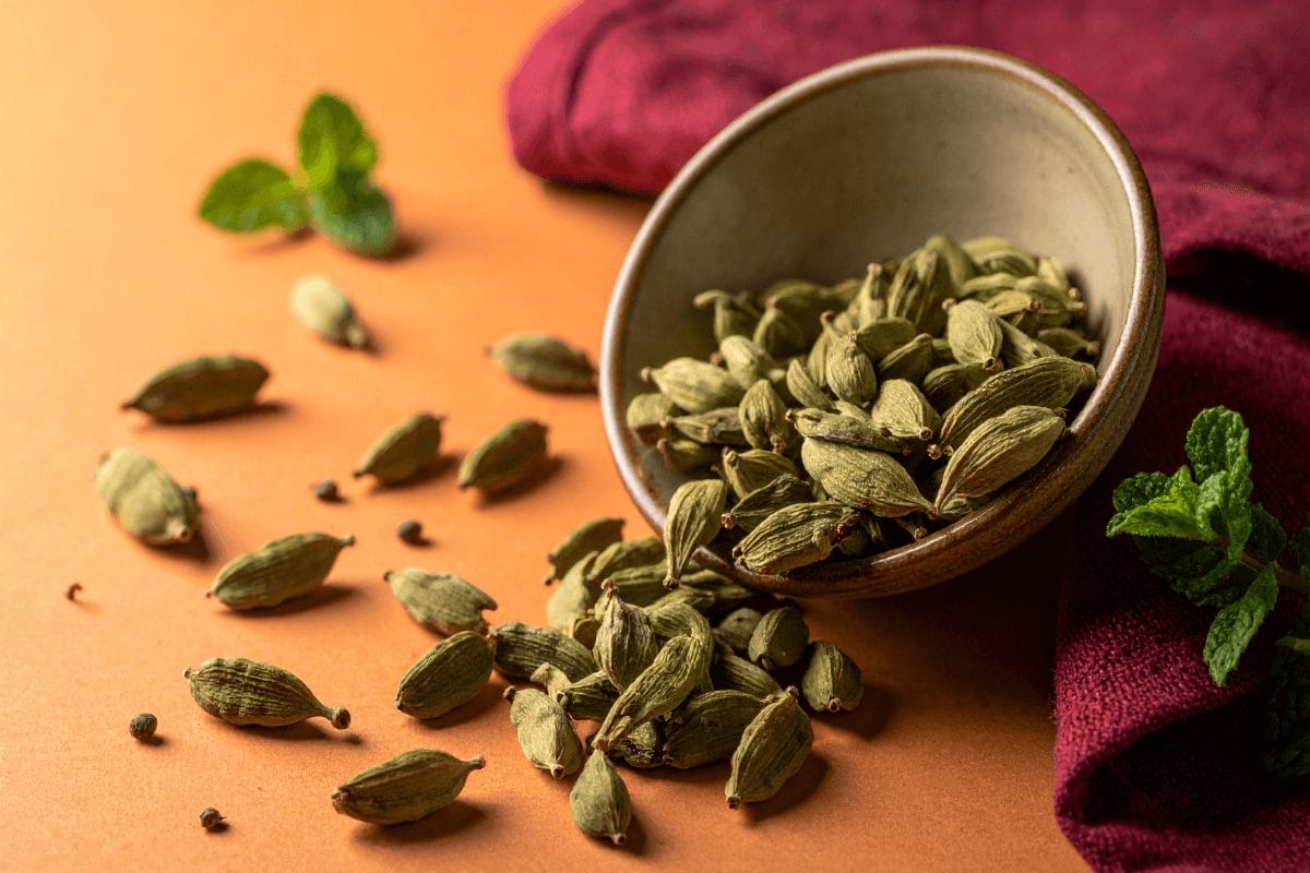 Green cardamom pods spilling from bowl on pastel orange with burgundy linen – cardamom digestive herb.