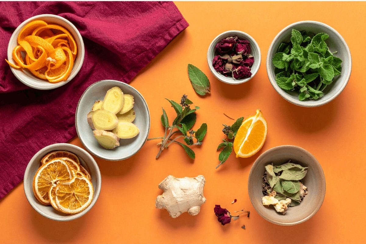 Flat lay of Ayurvedic kitchen herbs and spices — dried orange slices, tulsi leaves, ginger, mint, orange peel, and rose — styled on pastel orange with burgundy napkins, symbolizing herbal alchemy and everyday healing in the kitchen.