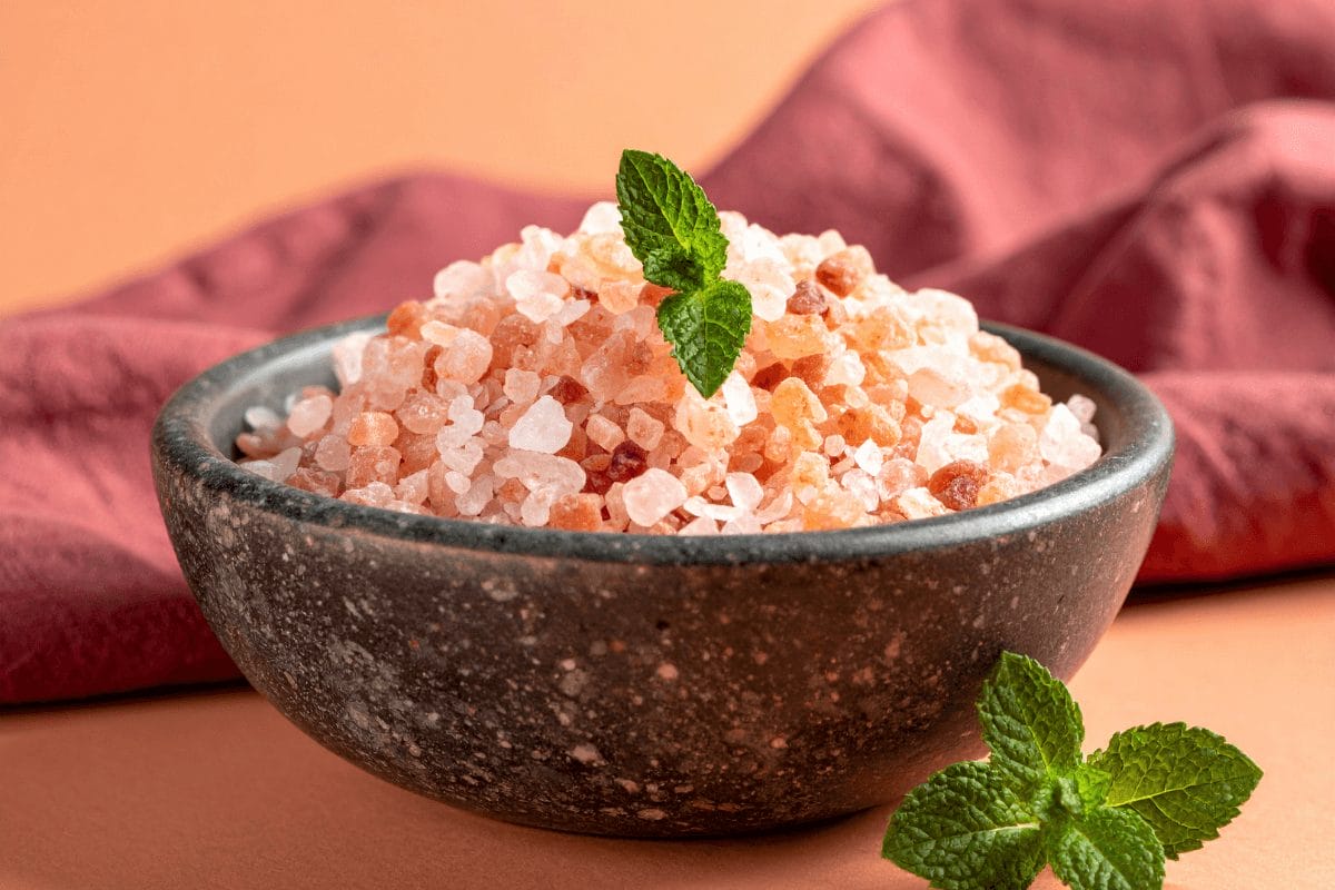 Pink Himalayan salt crystals in rustic ceramic bowl on pastel orange with burgundy linen – mineral-rich salt for digestion and balance.
